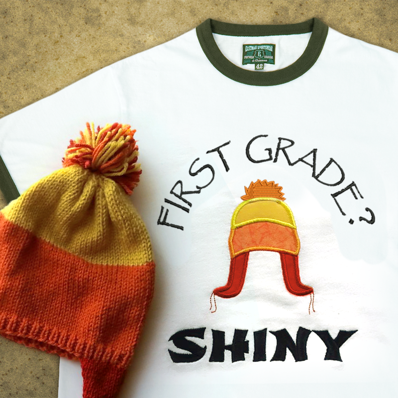 A ringer tee that is embroidered "First grade? Shiny" with an applique winter hat in orange, yellow, and red. A real hat in the same style sits on top.