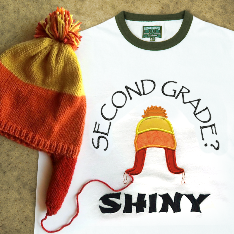 A ringer tee that is embroidered "Second grade? Shiny" with an applique winter hat in orange, yellow, and red. A real hat in the same style sits on top.