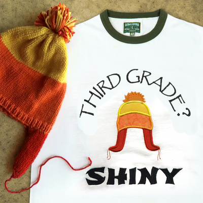 A ringer tee that is embroidered "Third grade? Shiny" with an applique winter hat in orange, yellow, and red. A real hat in the same style sits on top.