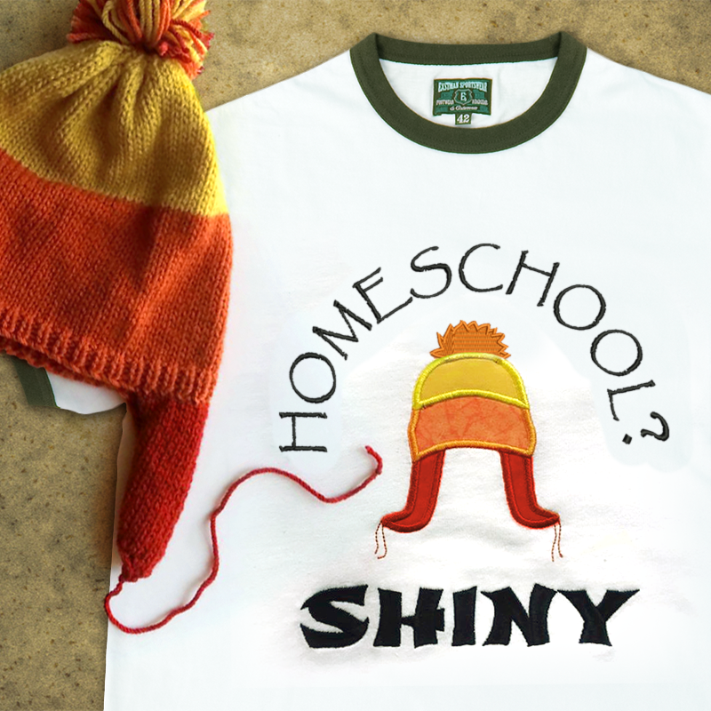 A ringer tee that is embroidered "Homeschool? Shiny" with an applique winter hat in orange, yellow, and red. A real hat in the same style sits on top.