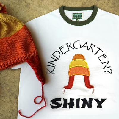 A ringer tee that is embroidered "Kindergarten? Shiny" with an applique winter hat in orange, yellow, and red. A real hat in the same style sits on top.