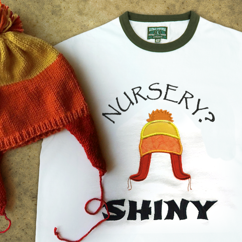 A ringer tee that is embroidered "Nursery? Shiny" with an applique winter hat in orange, yellow, and red. A real hat in the same style sits on top.