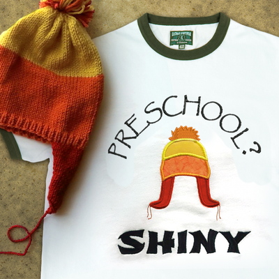 A ringer tee that is embroidered "Preschool? Shiny" with an applique winter hat in orange, yellow, and red. A real hat in the same style sits on top.