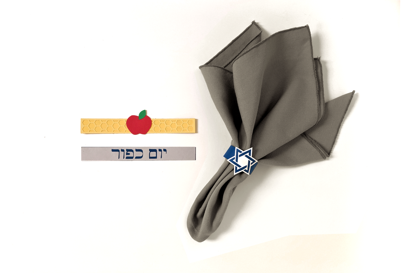 Grey napkin with a paper napkin ring that has a Star of David design. There are 2 more paper napkin rings laid flat nearby. One has an apple and the band looks like honeycomb. The other says "Yom Kippur" in Hebrew letters.