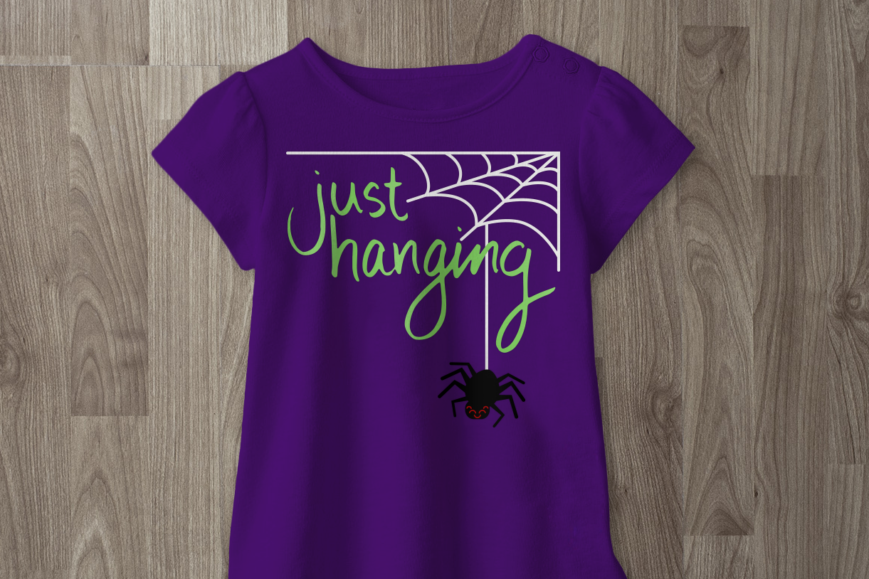 Purple tee with a spiderweb with a cute smiling spider and the text "just hanging."