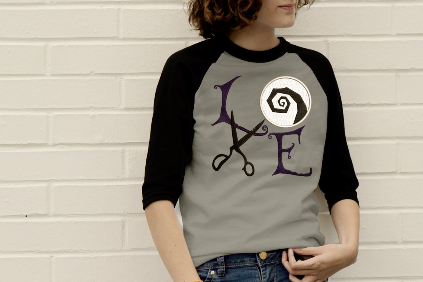 White-presenting teen girl wears a raglan tee with the applique word "LOVE" set on a square. The letters are in gothic style. In place of the O is a circle with a rough swirl embroidery. In place of the V are a pair of old scissors.