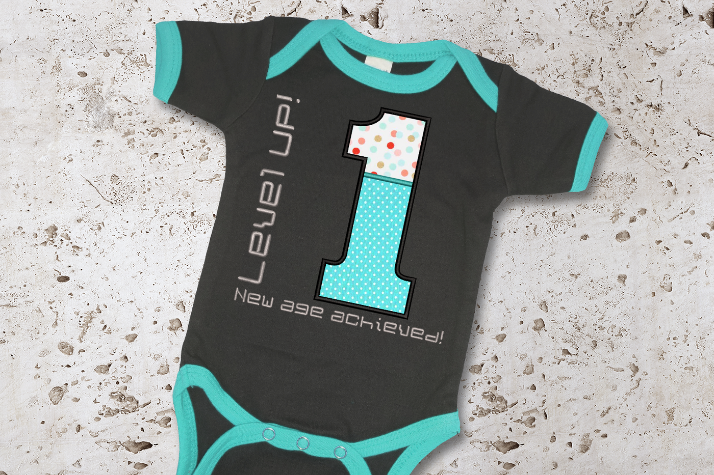 Baby onesie with a large applique 1. Around the 1 is embroidered "Level UP! New age achieved!"