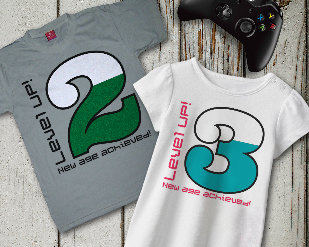 Two folded shirts. Each says "Level UP! New age achieved!" On has a large number 2, the other has a large number 3.