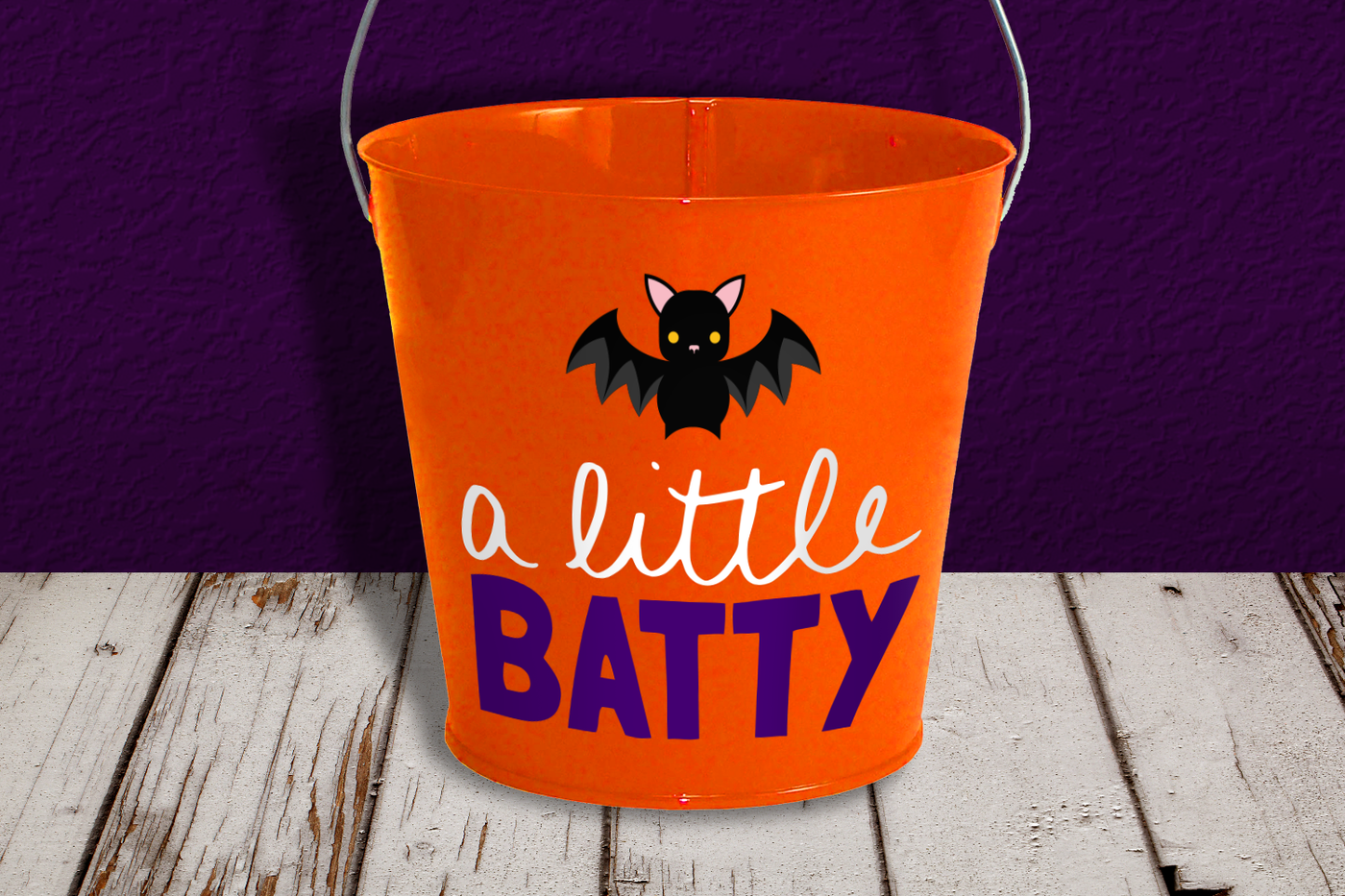 An orange pail sits on a wood floor with a purple wall behind it. There is a cute bat design and the words "a little batty"