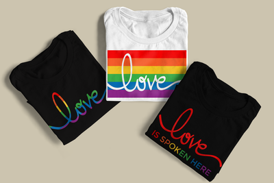 Three folded tees. One says "love" in rainbow stripes, another has rainbow stripes with the word "love" knocked out of it, and the last says "love is spoken here" in rainbow letters.