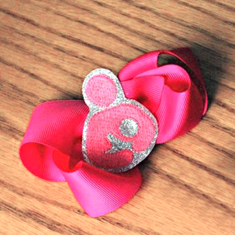 A hot pink bow sits on a wood surface. On the bow is a piece of glittery silver material with a pink breastfeeding symbol embroidered onto it.