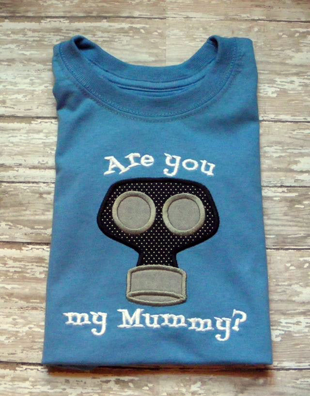 A folded blue shirt sits on a weathered wood background. Embroidered onto the shirt are the words "Are you my Mummy?" with an applique gas mask.