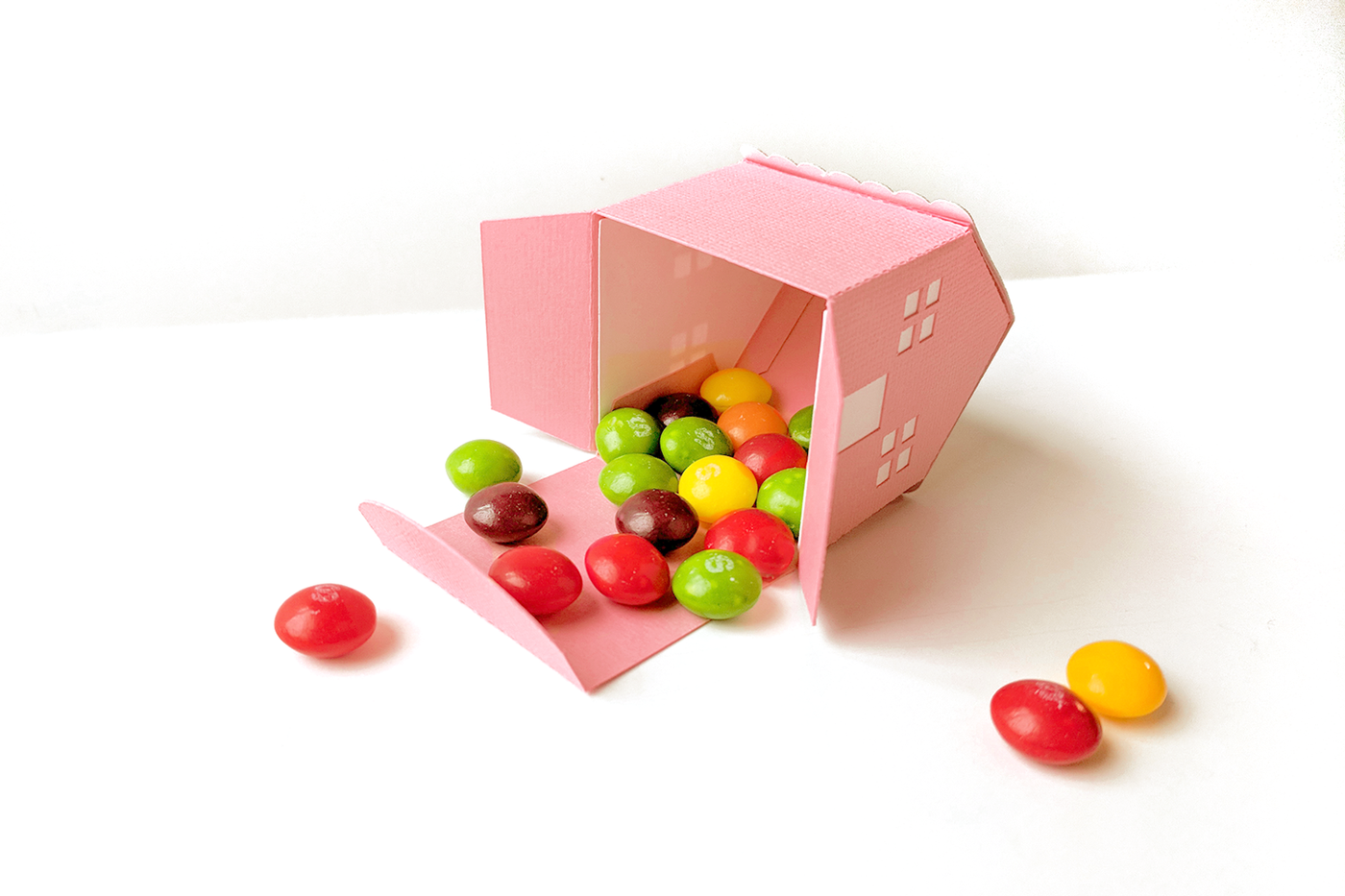 A 3D house box made of pink paper. The bottom of the box is open and spilling out Skittles candies.