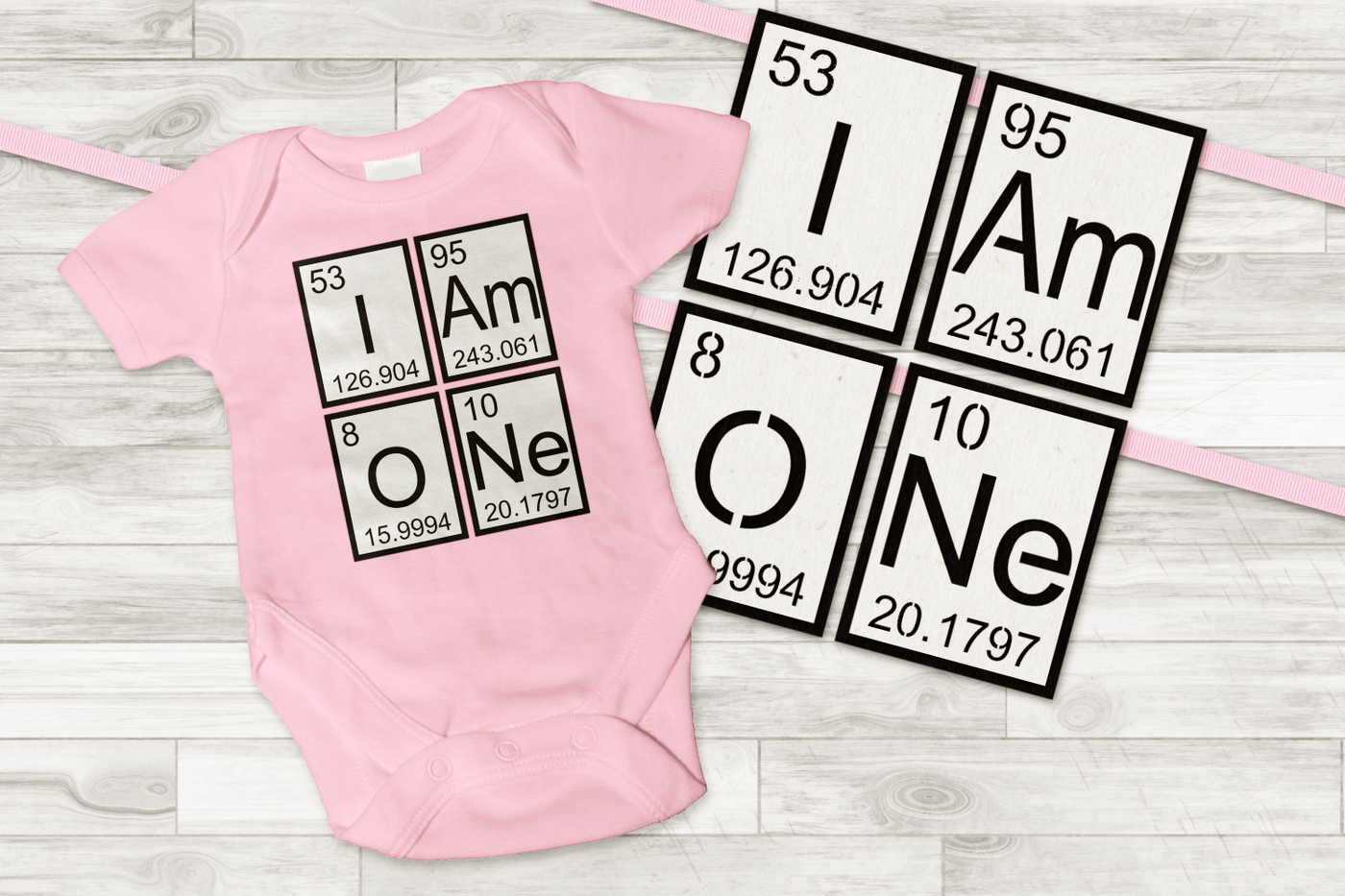 A pink baby onesie that says "I Am ONe" made out of periodic elements. The same periodic elements are used to make a banner.