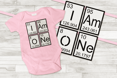 A pink baby onesie that says "I Am ONe" made out of periodic elements. The same periodic elements are used to make a banner.