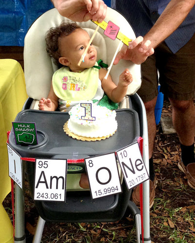 A black baby in a high chair has a cake in front of her. On the high chair, is a banner that says "I Am ONe" made out of periodic element symbols.