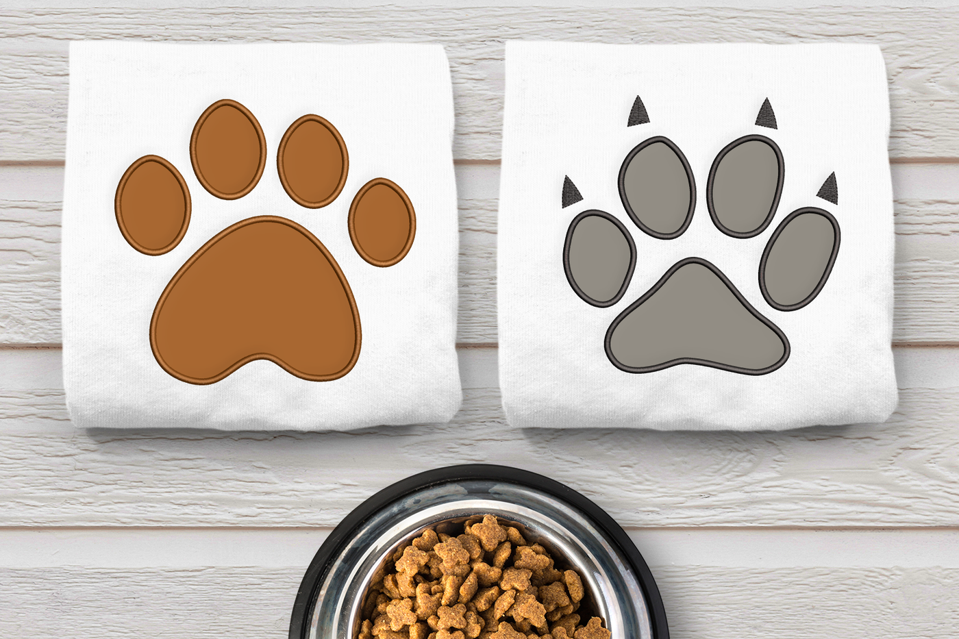 Dog and cat paw applique embroidery designs. Shown stitched onto white folded fabric. The fabric is on a wooden surface with a pet bowl nearby.