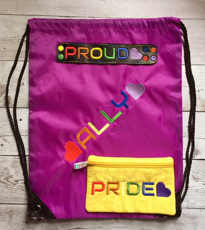 Drawstring bag with the word "Ally" and a heart at each side embroidered in rainbow colors. Black faux leather bracelet embroidered with "PROUD" with a heart in rainbow colors. Yellow zippered bag with the embroidered word "PRIDE" and a heart in rainbow colors.