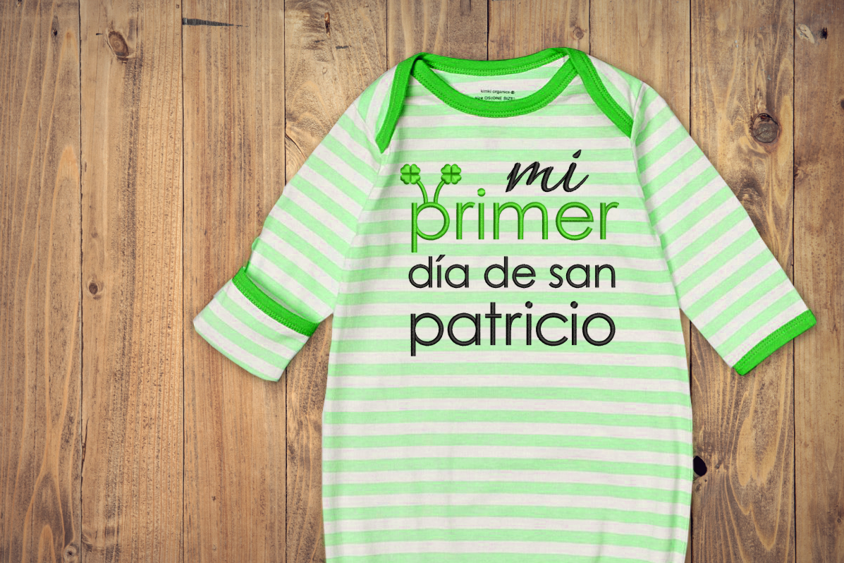 Green and white striped baby sleep gown on a wood background. Embroidered onto the gown is "mi primer dia de san patricio." The first P has clover antennae. 