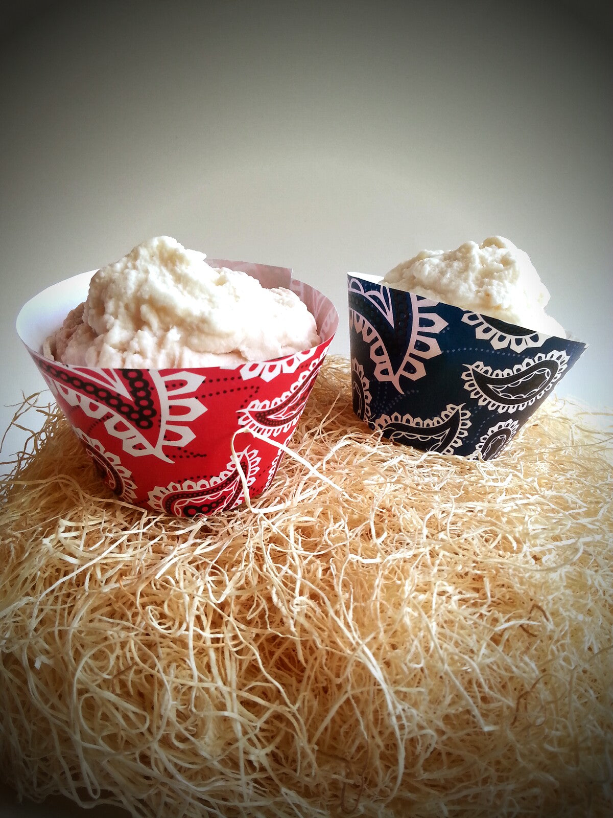 Two vanilla frosted cupcakes sit on a pile of hay. They have cupcake wrappers with flat tops resembling bandanna fabric. The left is red with black and white print, and the right is blue  with white and black print.