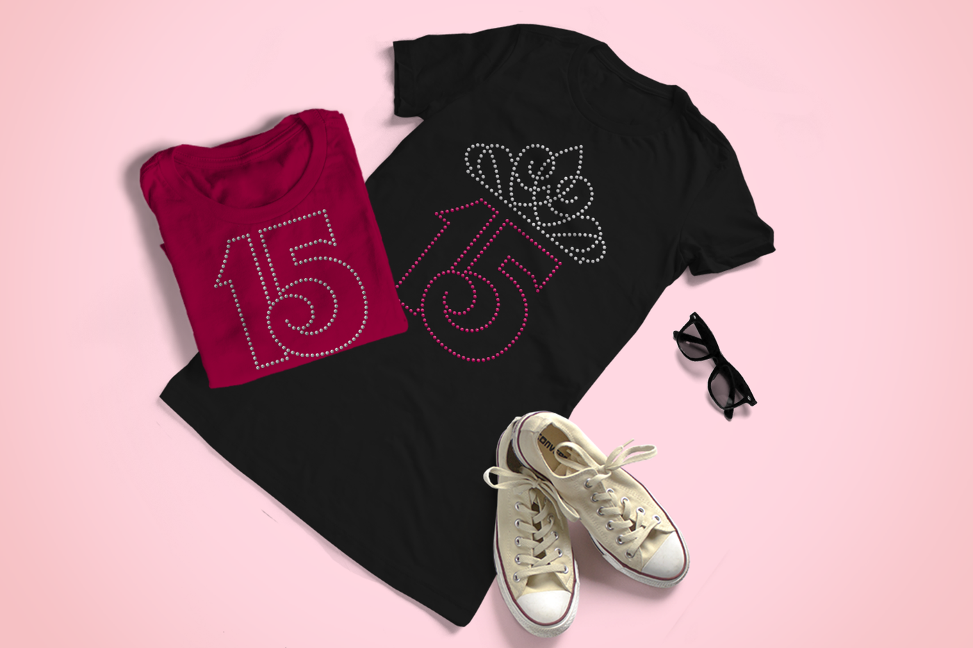 A black tee and a dark pink folded tee sit on a pale pink background, along with a pair of ivory Converse and sunglasses. The pink tee has a large number 15 in clear rhinestones. The black tee has the same 15 in hot pink rhinestones with a tiara in clear rhinestones above.