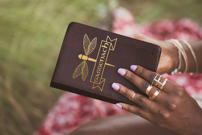 Black woman holds a notebook with a design of a Dragonfly with banner that says "Sassenach."
