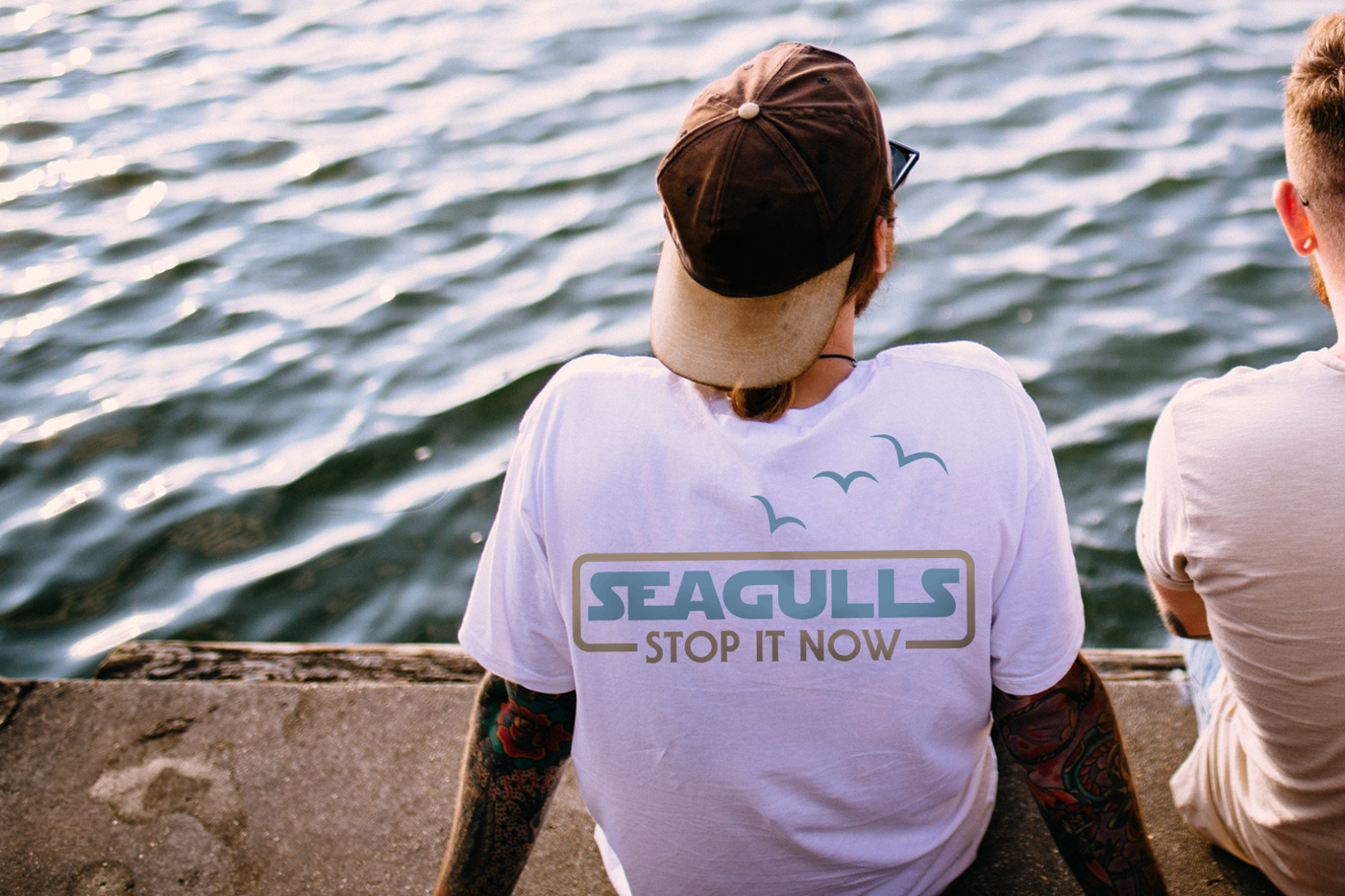 Two white men sit on a dock and are seen from the back. One man has a design on the back of his shirt that says "Seagulls stop it now" with 3 birds flying