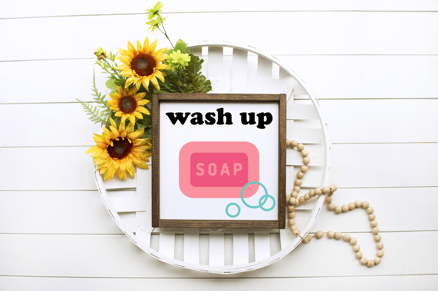 A decorative wicker plate with sunflowers sits on a white paneled backdrop. On to is a square wooden frame with a design that says "wash up" with a bar of pink soap and bubbles.