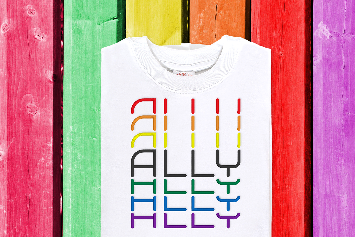 Ally embroidery design with stacked text. Shown on a white shirt laying on a rainbow painted wood background.