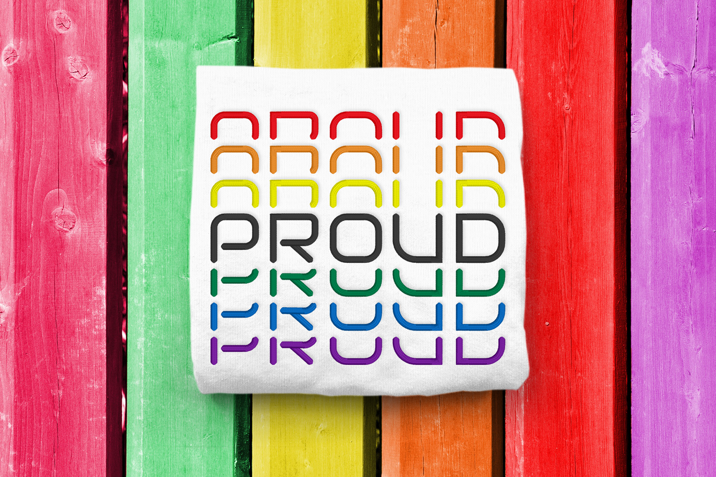 Stacked Proud embroidery design shown stitched on a white shirt on a rainbow painted wood background.