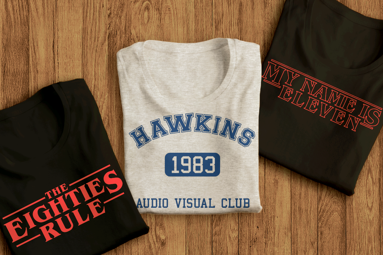 Three folded shirts on a wood panel background. Left shirt says "The Eighties Rule", middle shirt says "1983" with "Hawkins Audi Visual Club" around it, and the right shirt says "My name is Eleven."