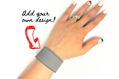 The hand of a white-presenting Latinx woman wearing a grey faux leather bracelet. An arrow points to the bracelet and says "Add your own design!"