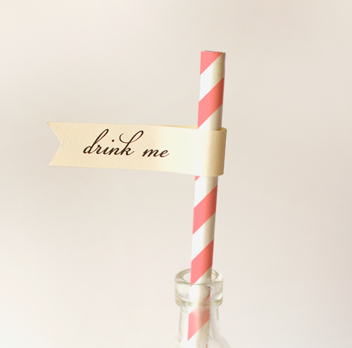 A pink and white striped straw with an ivory straw flag that says "drink me."