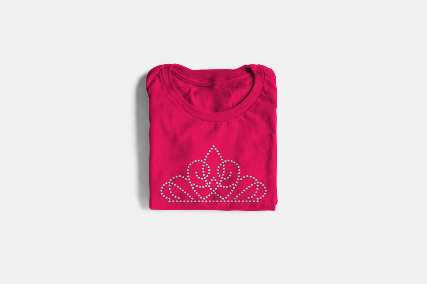 A hot pink folded tee on a light grey background. The tee has a tiara design in clear rhinestones.