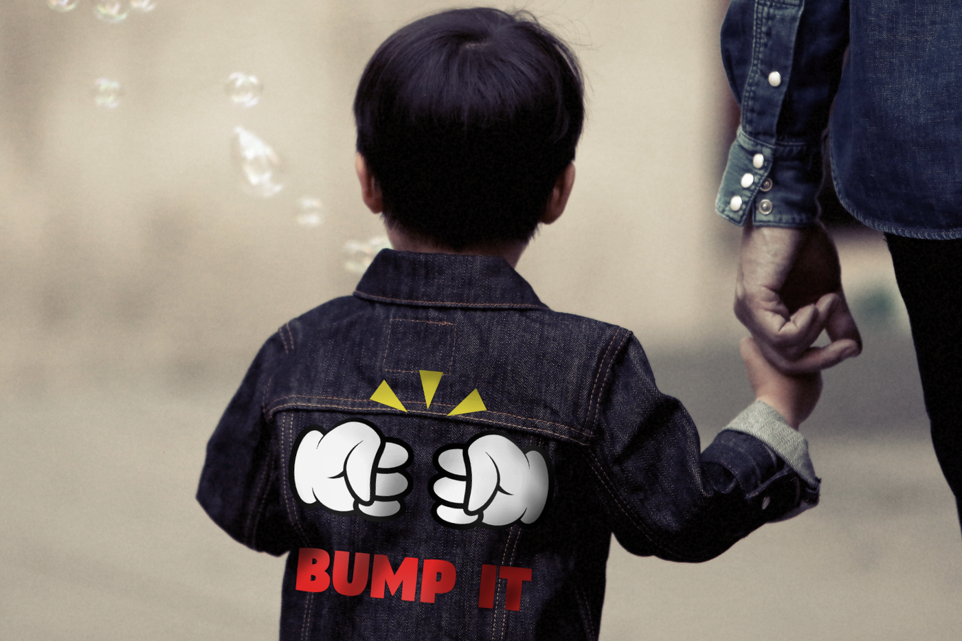An Asian toddler holds the hand of an adult and is seen from behind. He wears a denim jacket with a design of two white gloved cartoon fists doing a fist bump. The text below says "bump it" and above are 3 yellow triangles.