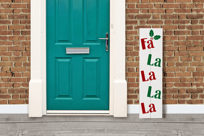 A door with a vertical sign leaning next to it. The sign has holly and says "Fa La La La La."