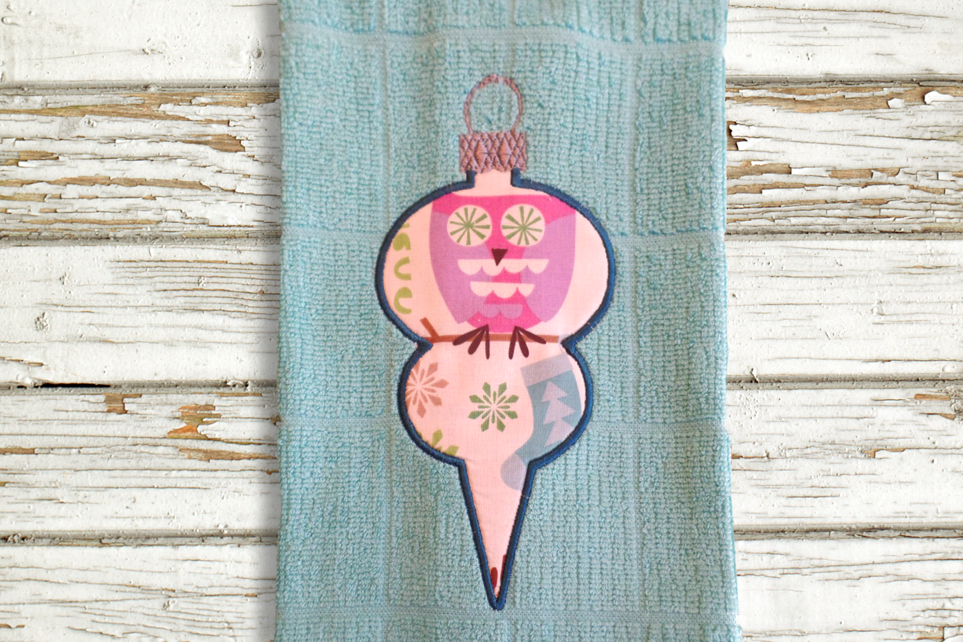 An aqua tea towel lays on a weathered painted wood surface. Appliqued onto the towel is an old fashioned Christmas ornament done in a retro owl holiday fabric.