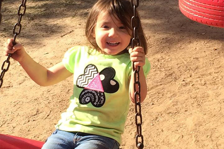 A little girl is on a swing, wearing a bright green shirt. The shirt has an applique heart an triangle design with black, white, and pink applique fabric.