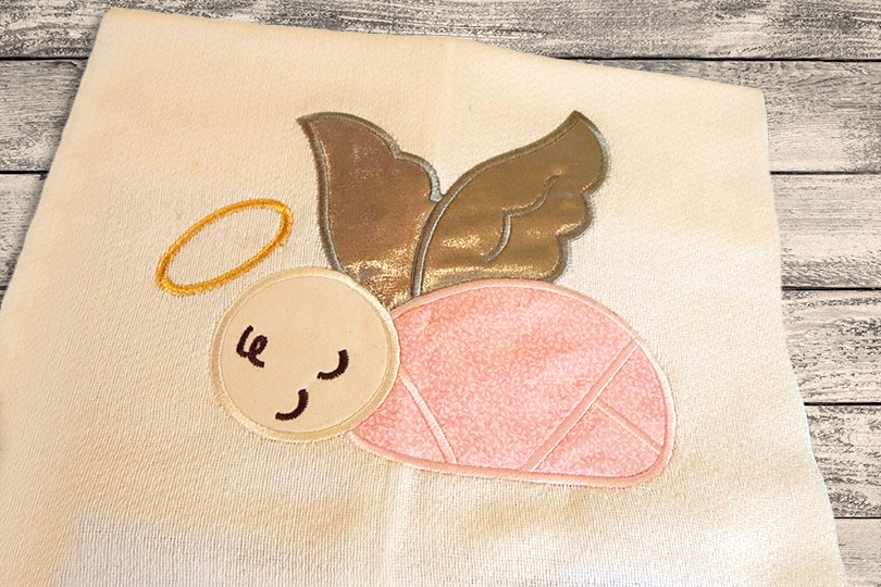 A square ivory piece of fabric lays on a weathered wood background. Appliqued onto the fabric is a baby in pink fabric bunting with peach skin, a halo, and gold wings.