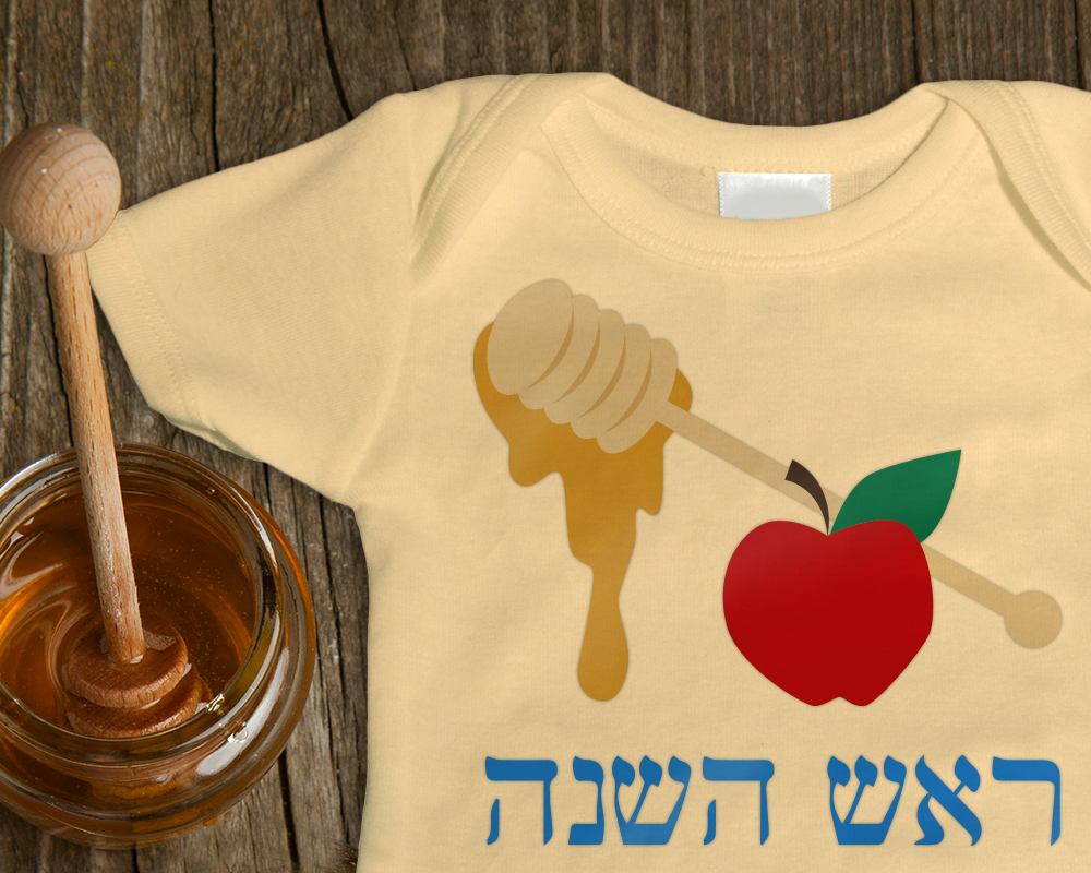A pale yellow baby onesie with a design of a honey wand dripping honey, an apple, and blue Hebrew letters. To the left of the shirt is a pot of honey.