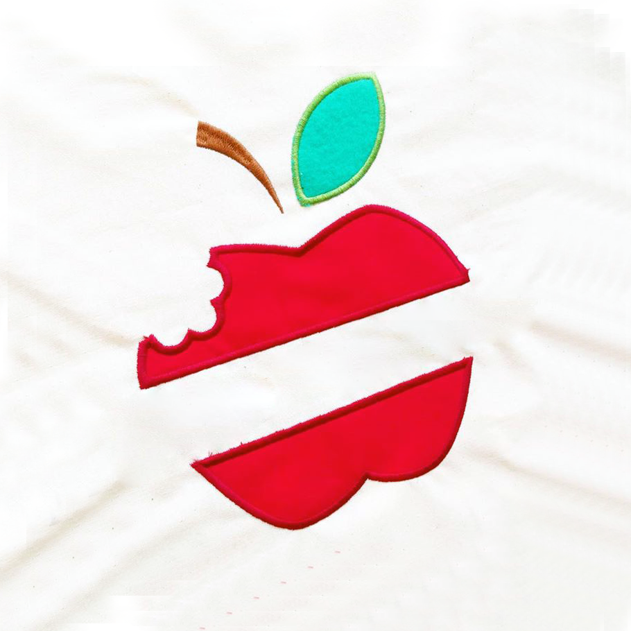 An apple with a bit out of it and split in the middle is appliqued onto a piece of white fabric.