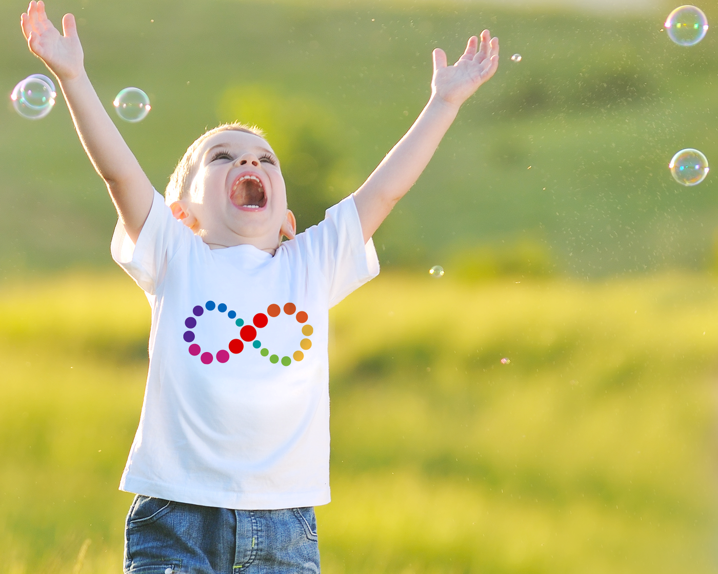 A laughing white child with arms raised chases bubbles outside. He is wearing a white shirt with an infinity symbol design made of circles in rainbow colors.