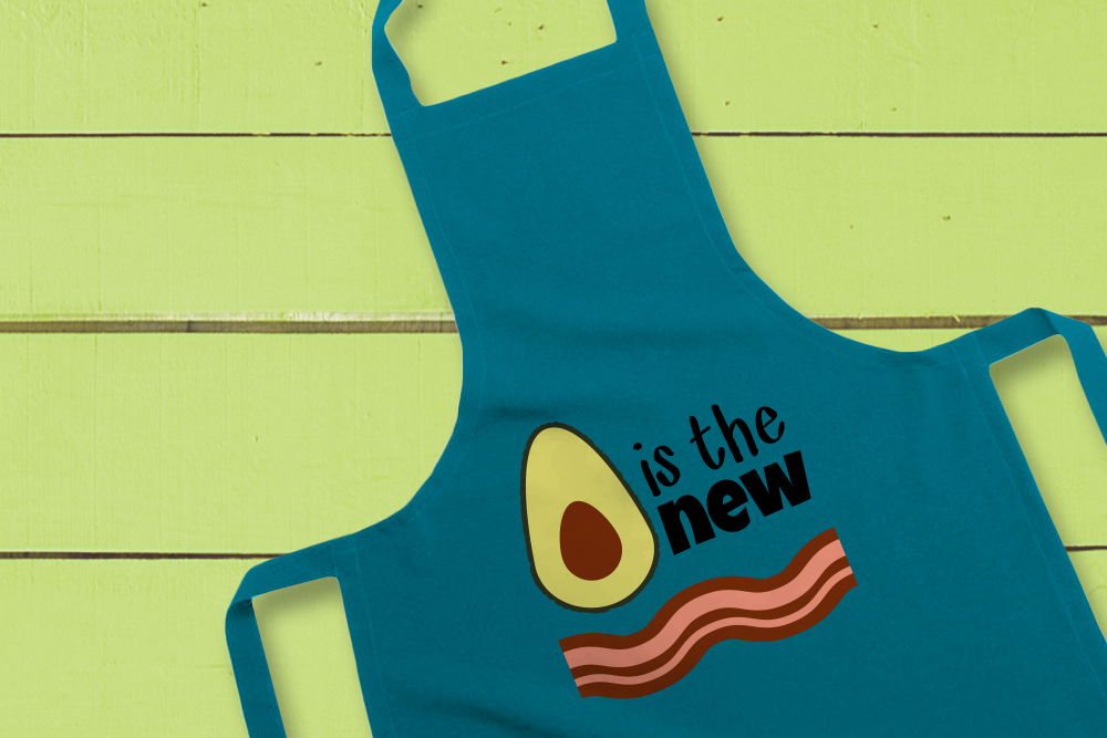A teal apron lays against a bright green painted wood backdrop. The apron has an image of an avocado and a strip of bacon, to make a phrase that reads "Avocado is the new bacon."