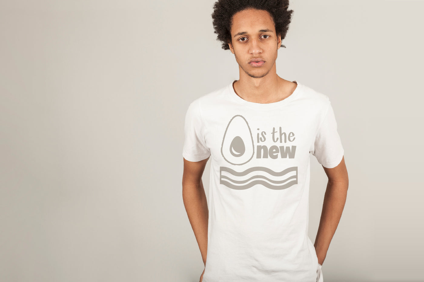 A black man wears a white tee with an image of an avocado and some bacon to be read as "Avocado is the new bacon." The design is in grey. He stands against a pale grey wall.