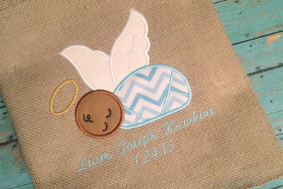 A piece of burlap lays on a weathered aqua painted wood background. Appliqued onto the burlap is a bundled sleeping baby with dark tan skin, a halo, wings, and blue chevron fabric for the bunting. Below the customer has added an embroidered name and date. (Text is not included in the design).