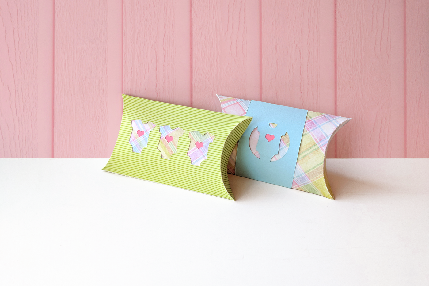 Two pillow boxes on a white surface with a pink paneled wall behind. The left box is green with cutouts of 3 pastel plaid baby onesies with pink hearts. The right box is pastel plaid with a blue bland that has the cutout of a baby onesie with a pink heart.