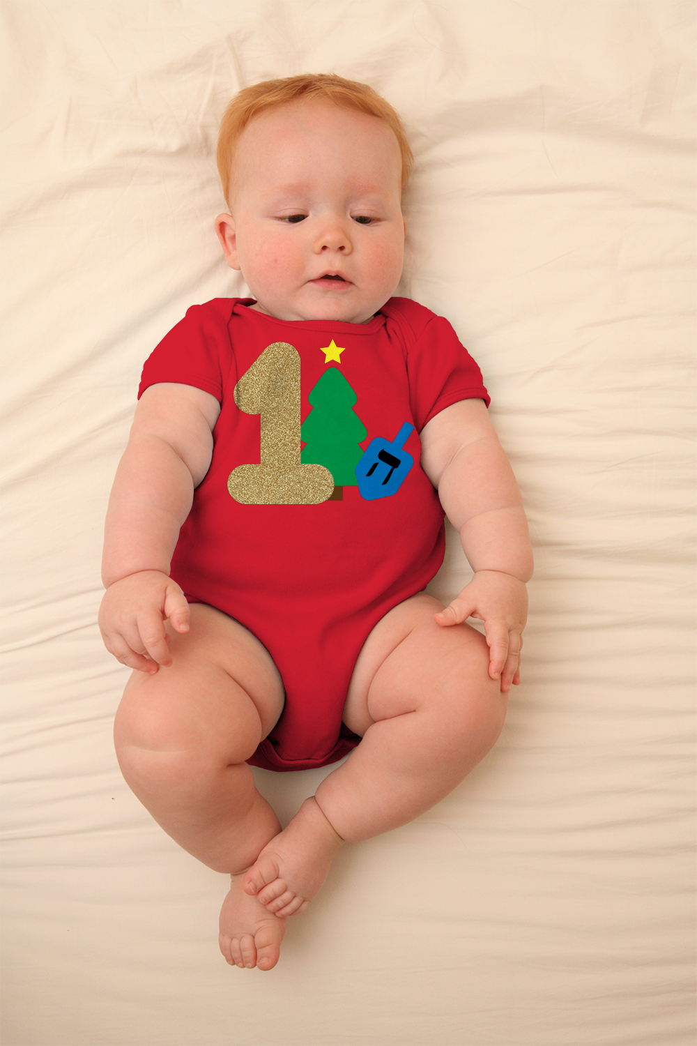 A white baby with a red onesie lays on a cream blanket. Their shirt has a large gold 1 with a Christmas tree and dreidel behind it.