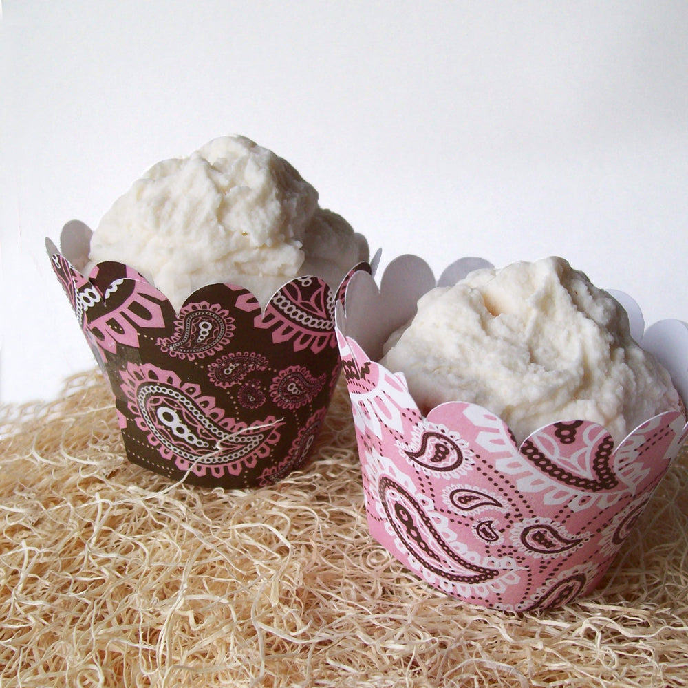 Two vanilla frosted cupcakes sit on a pile of hay. They have cupcake wrappers with scalloped tops resembling bandanna fabric. The left is brown with pink and white print, and the right is pink with white and brown print.