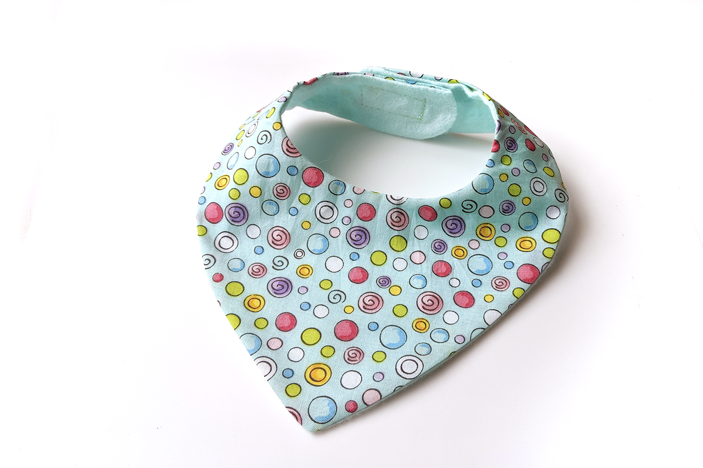 A bandanna style bib sits on a white background. The front bib fabric is aqua with bright dots in many colors and sizes outlined in black. The lining fabric is aqua flannel.