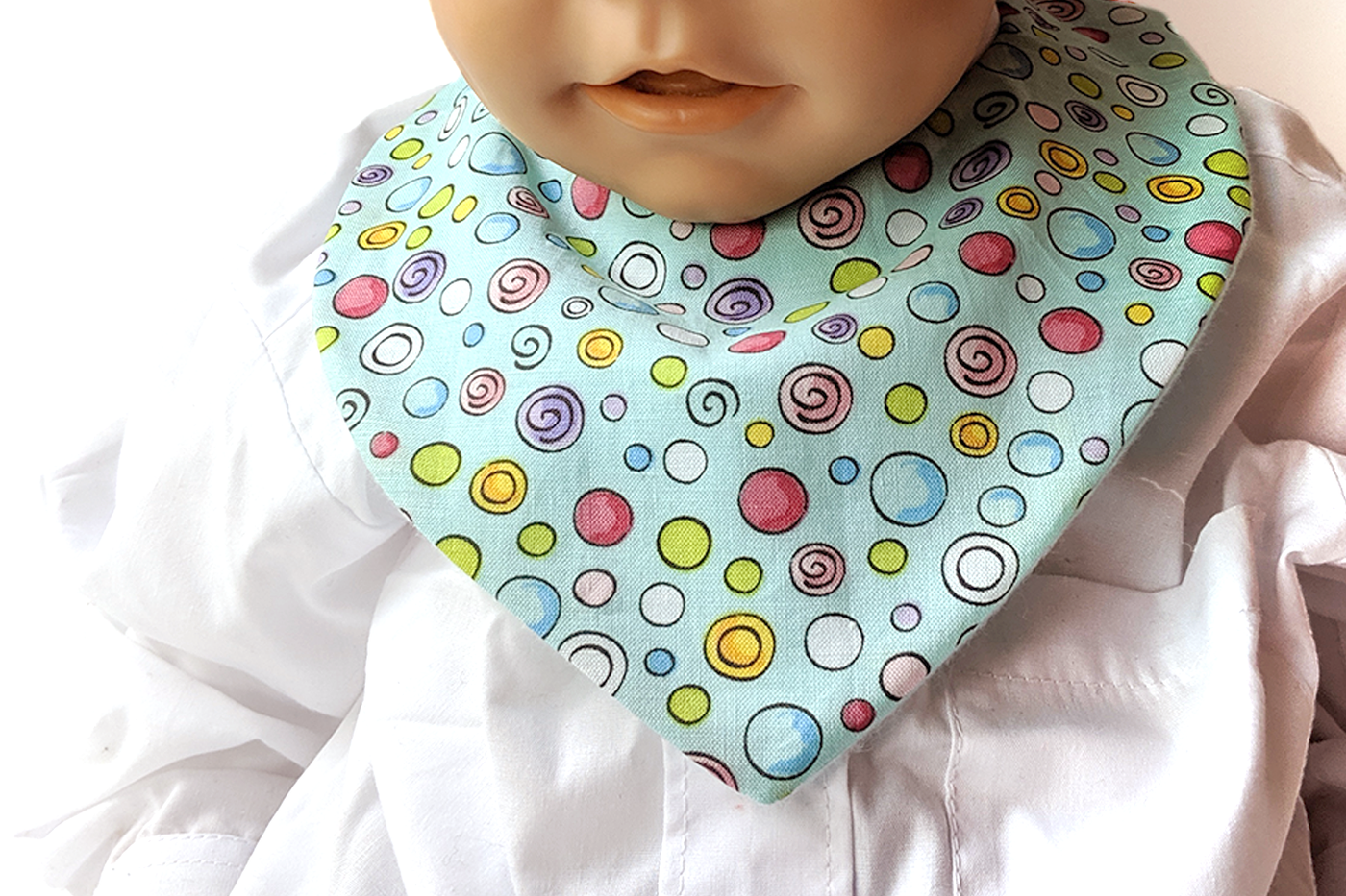 A white baby mannequin wears a white shirt and bandanna style bib. The bib fabric is aqua with bright dots in many colors and sizes outlined in black. 